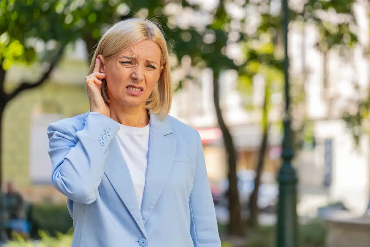 femme mature habillée en blazer bleu touchant son oreille avec une expression de douleur en extérieur